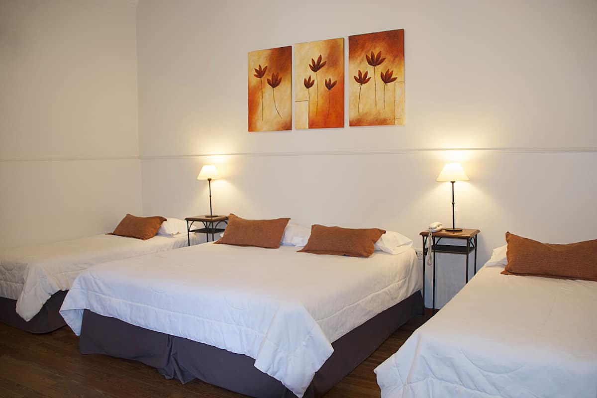 A hotel room with three single beds, each with a brown pillow and white bedding. There are bedside tables with lamps and a phone. The room has a wooden floor and white walls.