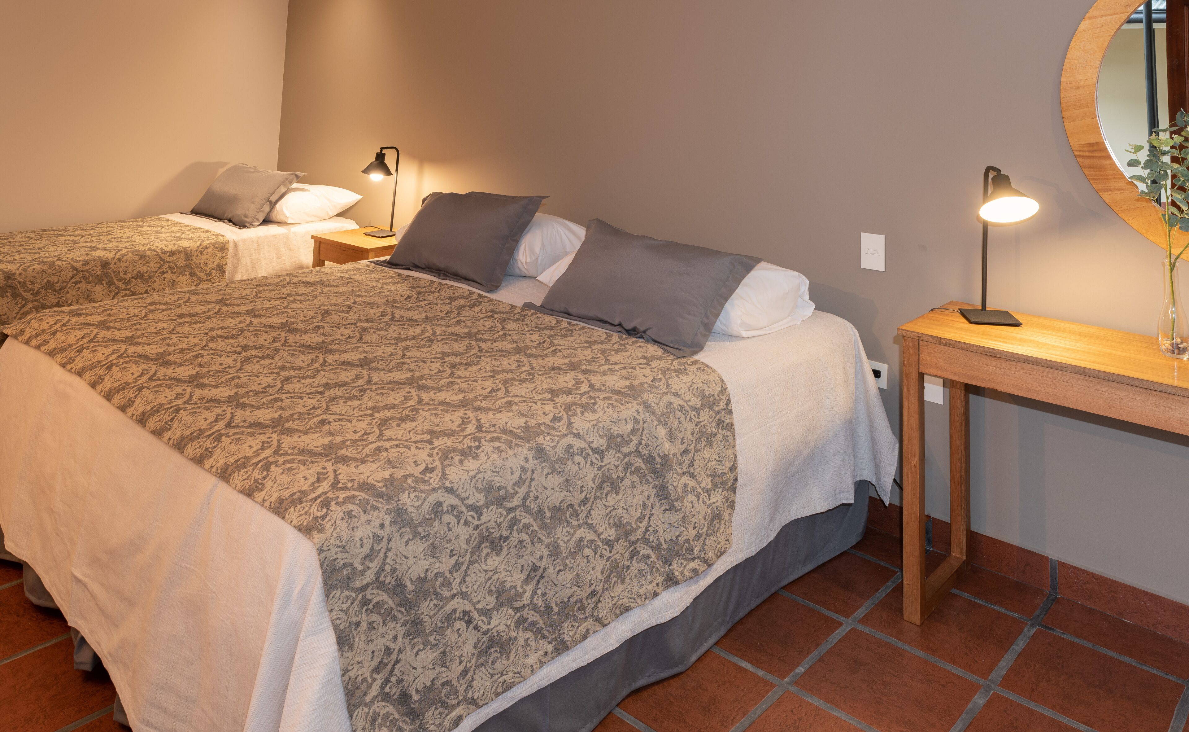 A bedroom with two beds, each with a patterned bedspread and pillows, a bedside table with a lamp, and a mirror on the wall.