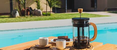 Uma mesa de madeira com uma cafeteira, xícaras e doces ao lado de uma piscina.