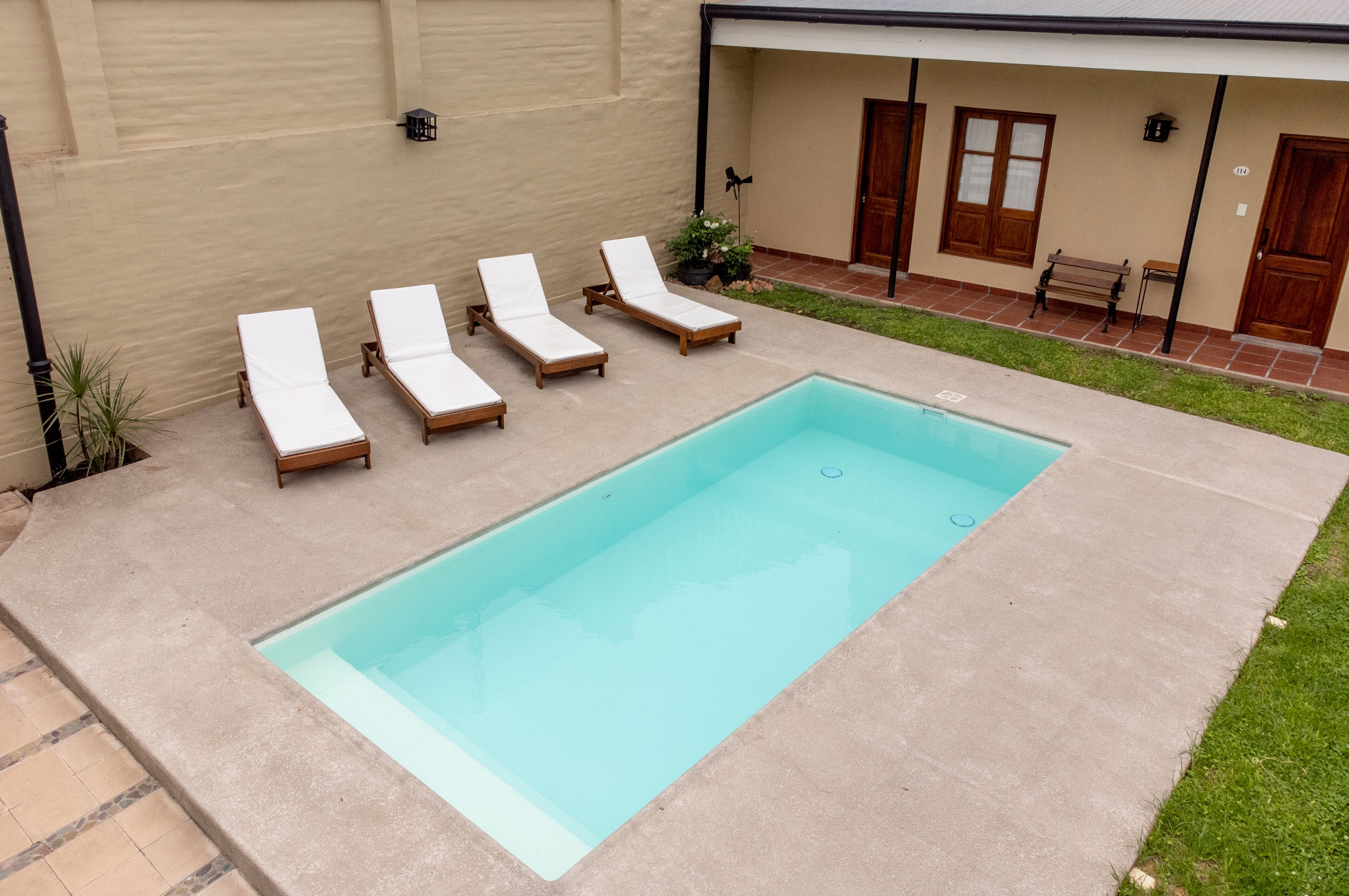 A rectangular swimming pool with four lounge chairs and a bench, surrounded by a concrete deck and a beige wall.