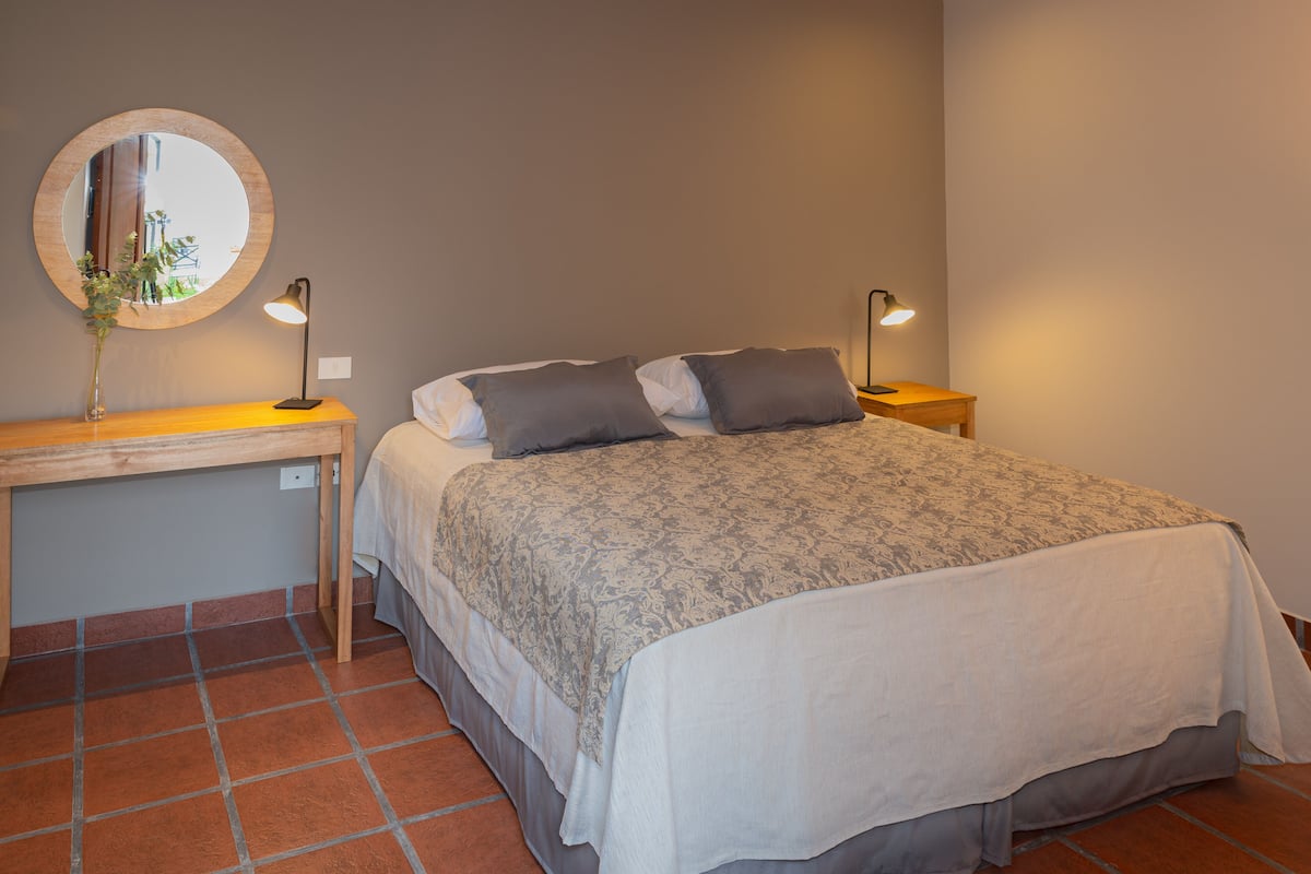 A bedroom with a bed, two bedside lamps, a round mirror, and a wooden desk with a vase.