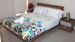 A neatly made bed with a floral bedspread, a bedside table with a lamp, and a painting of a coastal scene on the wall. - The New Waves (POINT COOK)