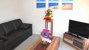 A living room with a black sofa, a wooden coffee table with a bouquet of flowers, a television on a wooden stand, and three beach paintings on the wall. - The New Waves (POINT COOK)