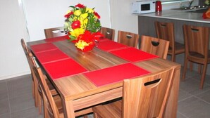 A wooden dining table with red placemats, surrounded by wooden chairs. A bouquet of flowers is placed on the table. - The New Waves (POINT COOK)