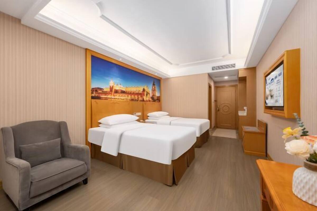 A hotel room with a bed, a chair, a desk, and a painting of a cityscape.