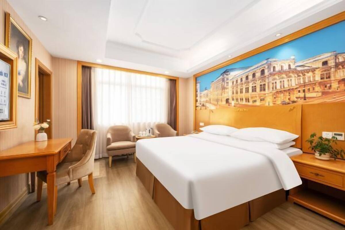 A hotel room with a bed, a desk, a chair, a nightstand, and a painting of a building on the wall.