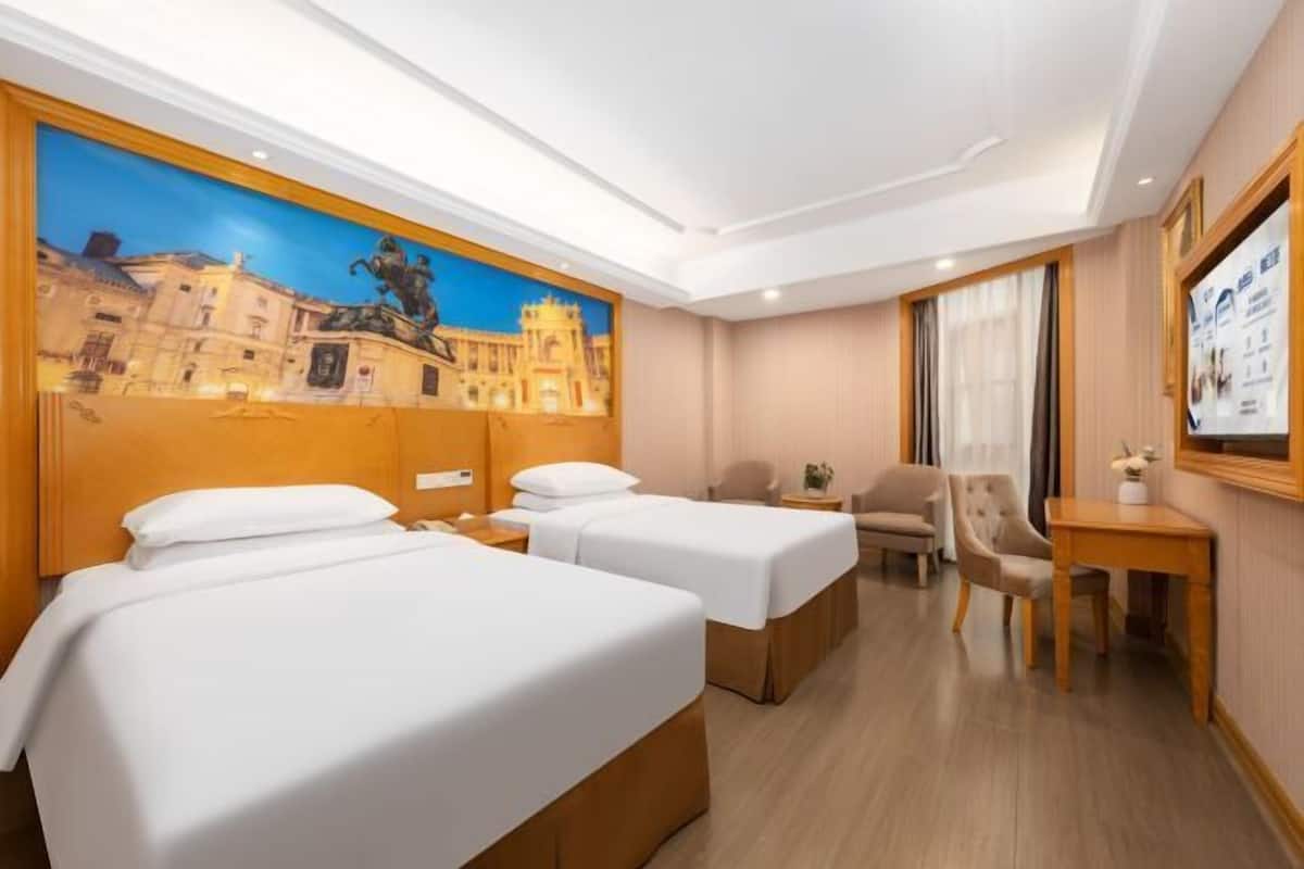 A hotel room with two beds, a desk, a chair, and a painting on the wall.