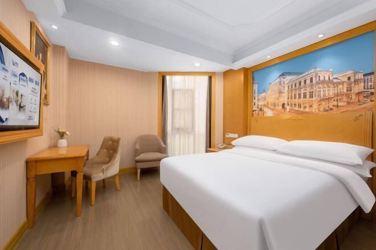 A hotel room with a bed, a desk, a chair, a window, and a painting on the wall.