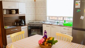 A compact kitchen with a stove, oven, sink, and a refrigerator. There is a dining table with a checkered tablecloth and a basket of flowers. - Cabañas Vaiora (Hanga Roa)