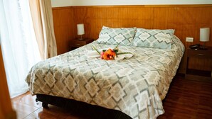 A neatly made bed with patterned bedding, a decorative flower arrangement, and wooden headboard and nightstands. - Cabañas Vaiora (Hanga Roa)