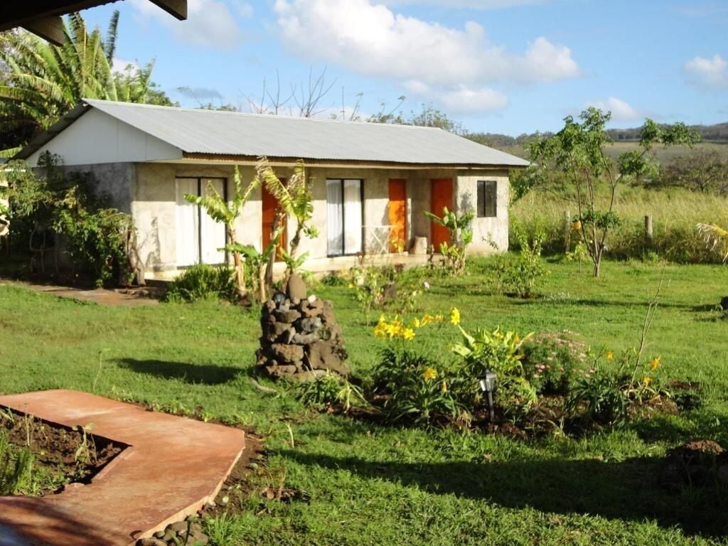 Una casa de un piso con techo inclinado, pared de piedra y un pequeño jardín con flores amarillas.