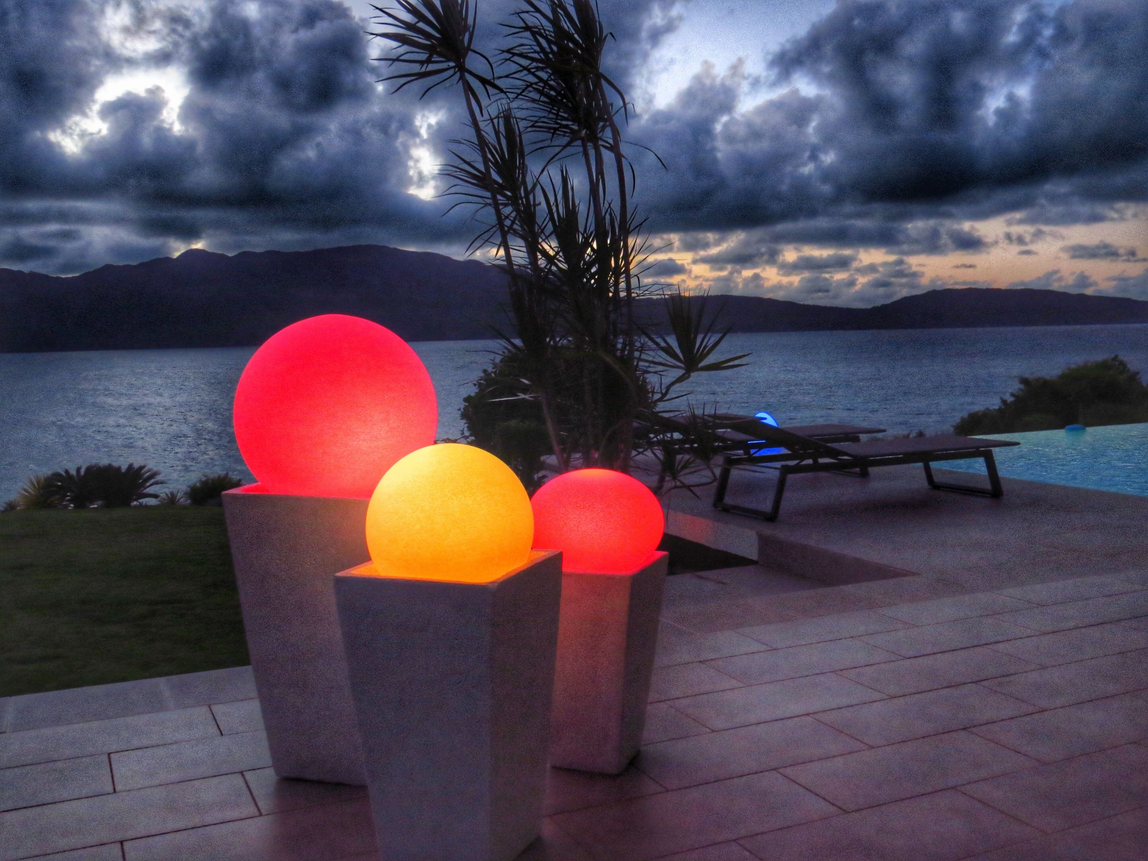 A poolside area with a view of the sea and mountains, featuring illuminated spherical lights and lounge chairs.
