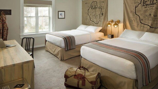Une chambre d’hôtel avec deux lits, un bureau, une chaise, une valise et une fenêtre.