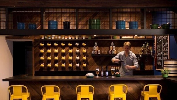 Un espace bar avec un barman, des tabourets jaunes et un mur rempli de bouteilles d’alcool.