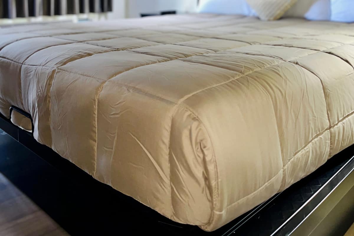 A bed with a quilted headboard, a bedside lamp, and a view of a window with blinds.
