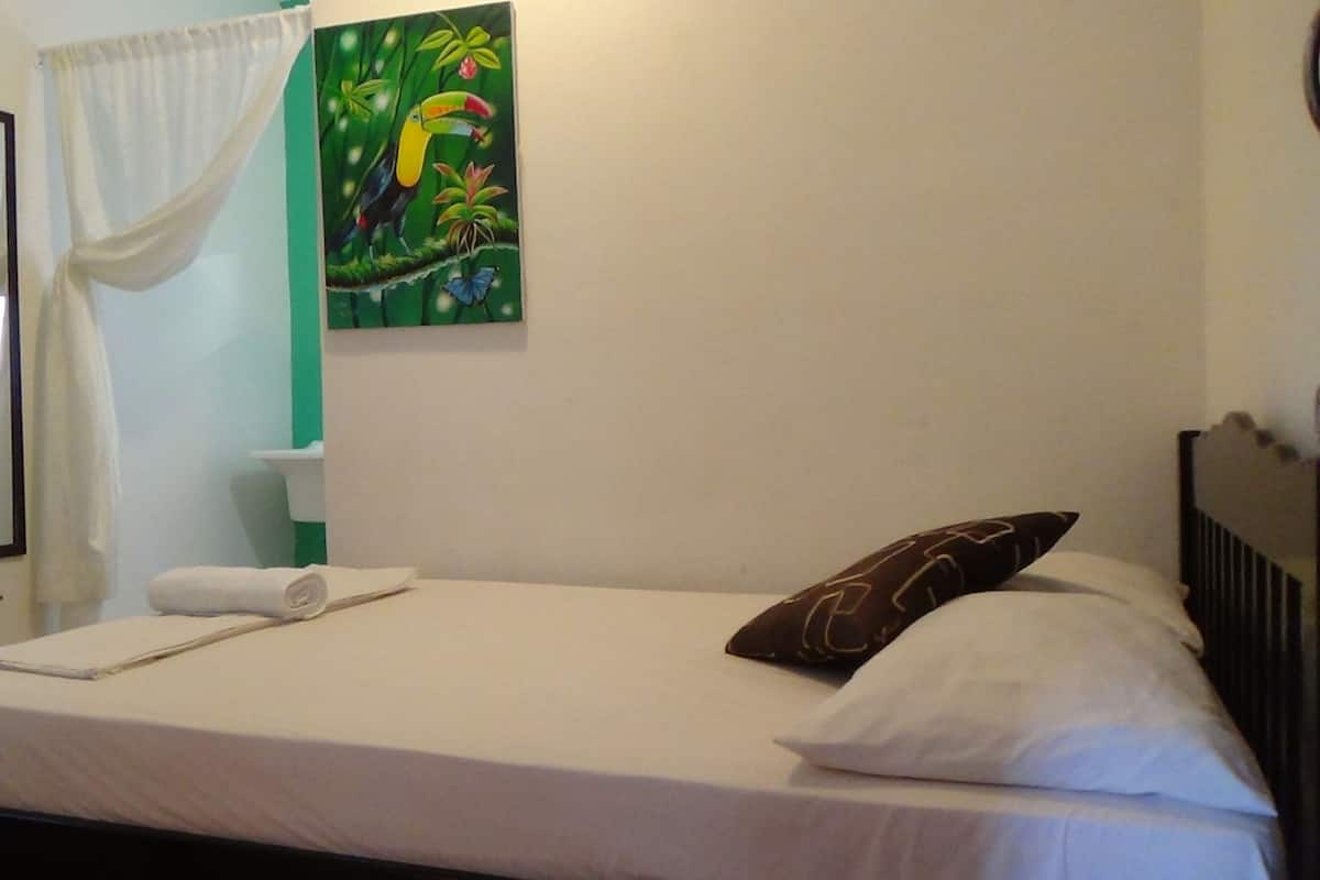 Un lit bien fait, avec du linge de lit blanc et un oreiller marron, un rideau blanc, un mur vert orné d’un tableau représentant un toucan, et une petite étagère avec des articles de toilette.