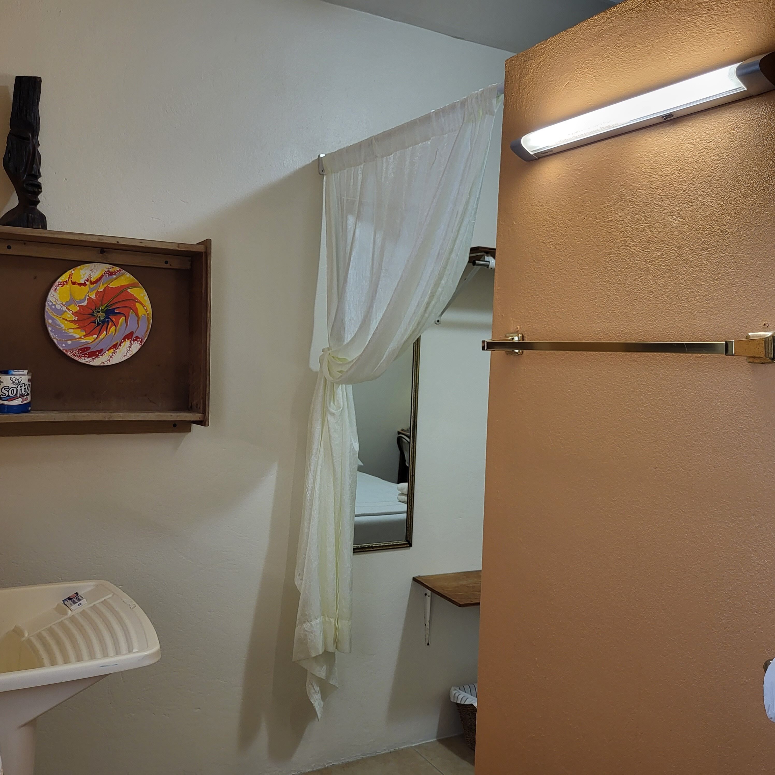 A bathroom with a white sink, a mirror, a shelf with a decorative plate, a white shower curtain, and a door with a light fixture.