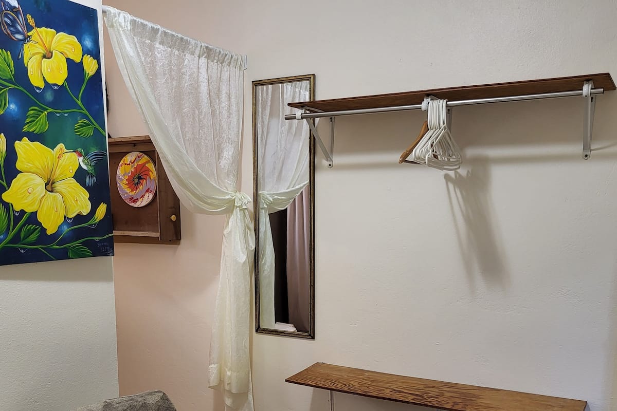 Un lit bien fait, avec du linge de lit blanc, un petit tabouret, une étagère en bois, un miroir et un tableau représentant des fleurs jaunes.