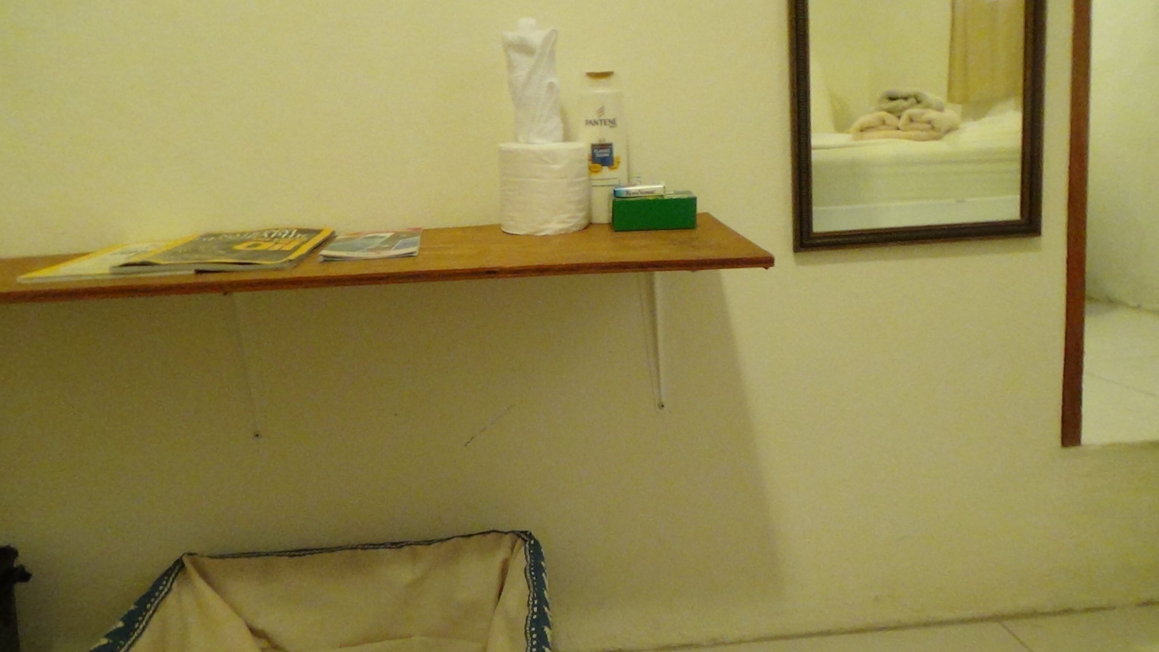 A small bathroom with a wooden shelf holding a tissue box, a bottle, and some magazines. A mirror reflects a bed with a white blanket.