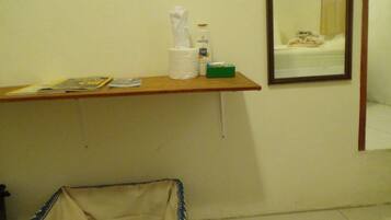 A small bathroom with a wooden shelf holding a tissue box, a bottle, and some magazines. A mirror reflects a bed with a white blanket.