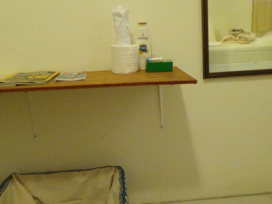 A small bathroom with a wooden shelf holding a tissue box, a bottle, and some magazines. A mirror reflects a bed with a white blanket.