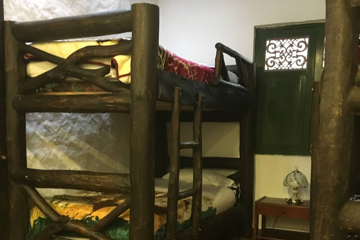 A bunk bed with a wooden frame and a patterned bedspread. There is a small table with a lamp next to a green door with decorative glass.