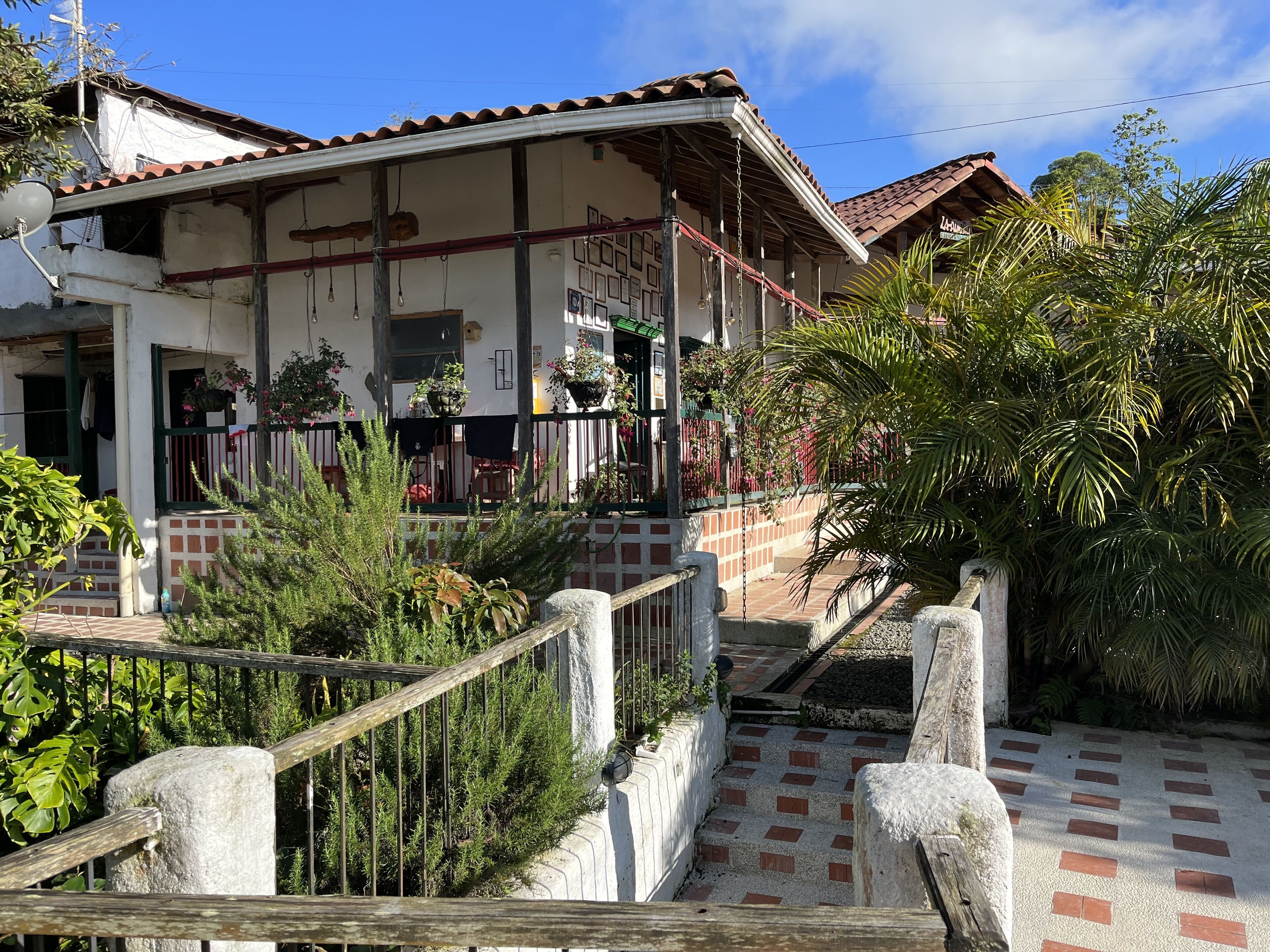 A two-story building with a tiled roof, a balcony with potted plants, and a paved entrance area with a stone railing.
