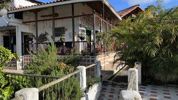 A two-story building with a tiled roof, a balcony with potted plants, and a paved entrance area with a stone railing.