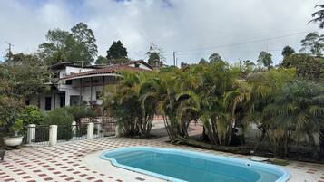 Una piscina con solárium de baldosas, rodeada de palmeras y una casa con techo rojo.