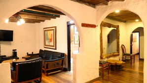 Uma sala de estar aconchegante com piso de madeira, um sofá de couro preto, uma poltrona de vime e uma mesa de centro de madeira. Há uma televisão de tela plana na parede e um quadro emoldurado sobre o sofá.