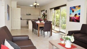 A modern living room with a dining area, a kitchen in the background, and a large window with blinds. - City Mews (GOWANBRAE)