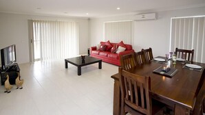 A modern living room with a red sofa, a dining table with chairs, a coffee table, and a flat-screen TV. - Villa Jemma (POINT COOK)
