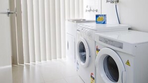 A laundry room with two front-loading washing machines, a drying machine, and a detergent dispenser on the wall. - Villa Jemma (POINT COOK)