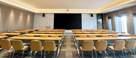 A conference room with rows of tables and chairs, a projector screen, and a large window.