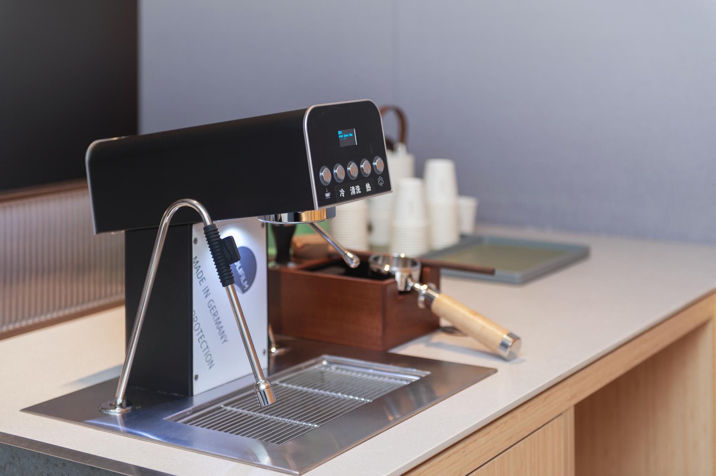 A coffee machine with a digital display, a metal drip tray, and a wooden container with cups.