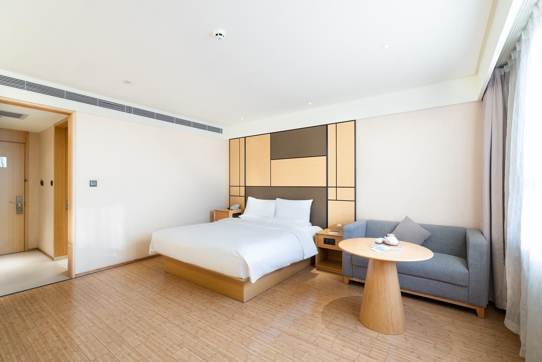 Una habitación de hotel moderna con una cama grande, una mesita de noche con lámpara, un sofá gris y una mesa redonda de madera.