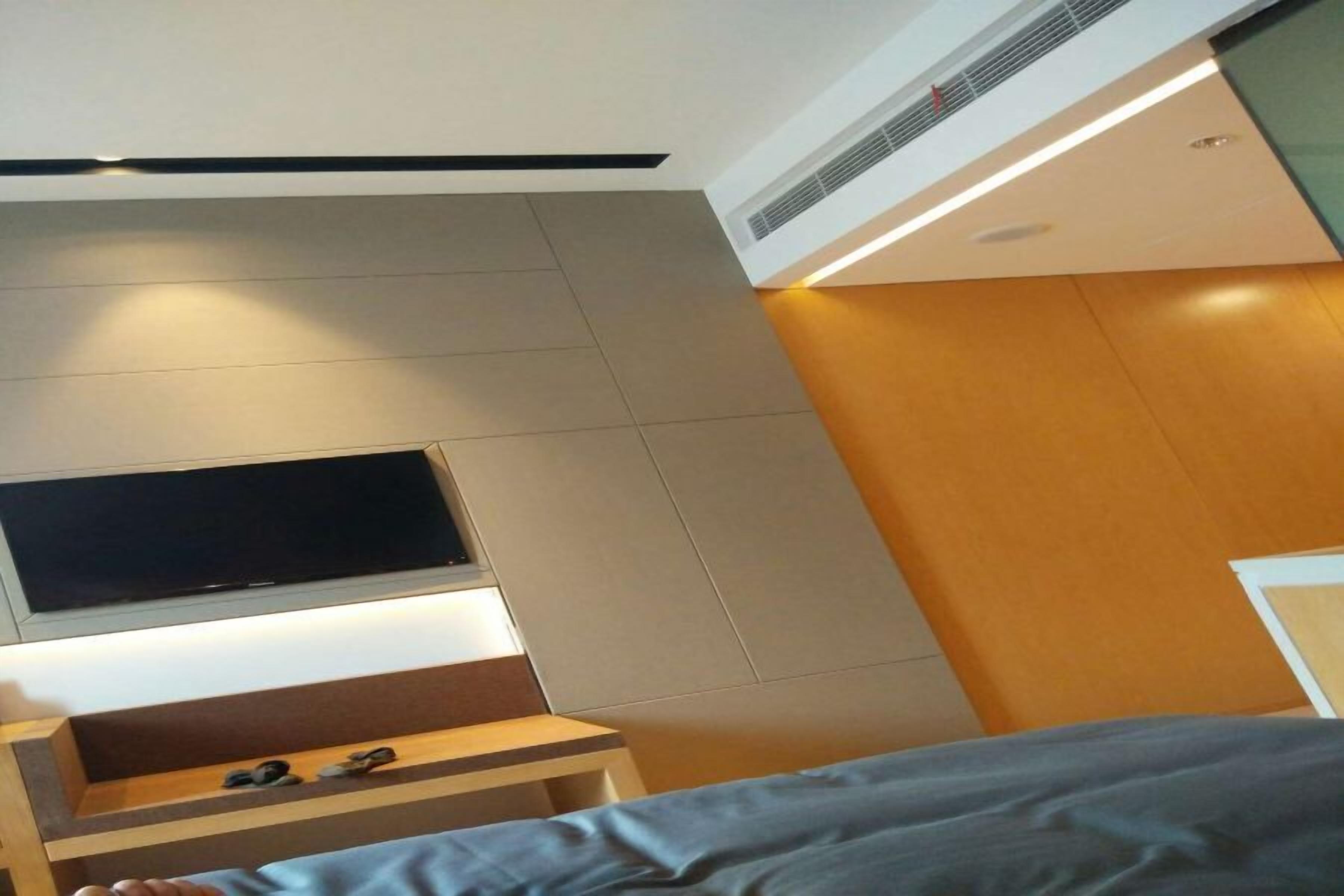 A modern hotel room with a flat-screen TV mounted on the wall, a wooden nightstand, and a bed with a grey bedspread.