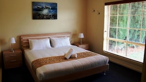 Een netjes opgemaakt bed met een beddeken met dessin, een opgerolde handdoek, twee bedlampen, een nachtkastje, een schilderij van boten aan de muur en een raam met jaloezieën.