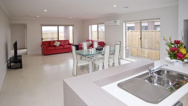 A modern living room with a red sofa, a glass dining table, and a kitchen area with a sink. - Villa Mystic (POINT COOK)