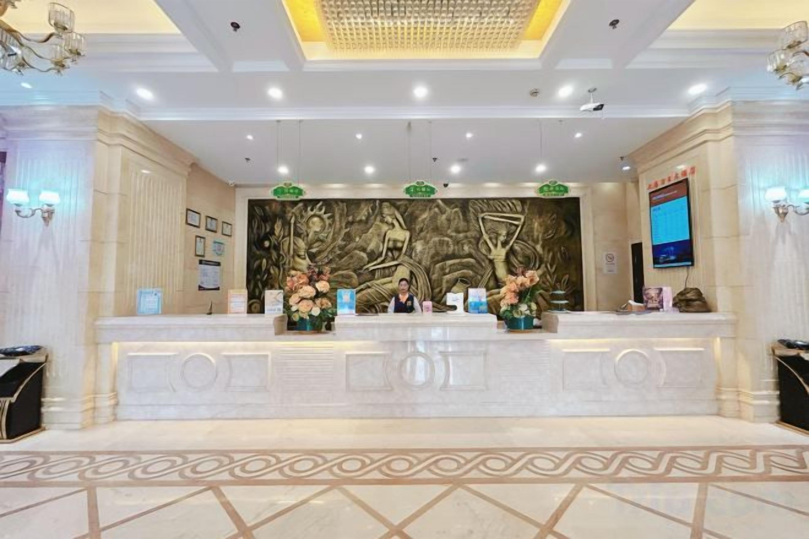A reception area with a marble counter, two receptionists, a large decorative wall, and a television screen.