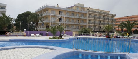 Zona de piscina con solárium de baldosas, sillas de descanso y un edificio con balcones al fondo.