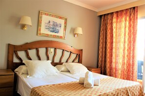 Een hotelkamer met een houten hoofdbord, twee bedlampen, een ingelijste foto aan de muur, een bed met witte lakens en een opgerolde handdoek op het bed.