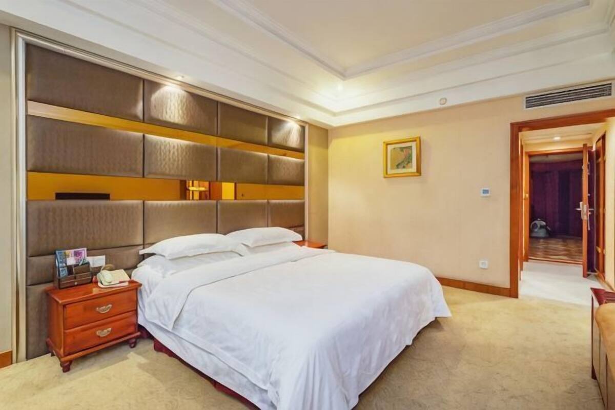 Une chambre d’hôtel avec un grand lit, une table de chevet en bois, une tête de lit dotée de compartiments de rangement et une porte donnant sur une autre pièce.