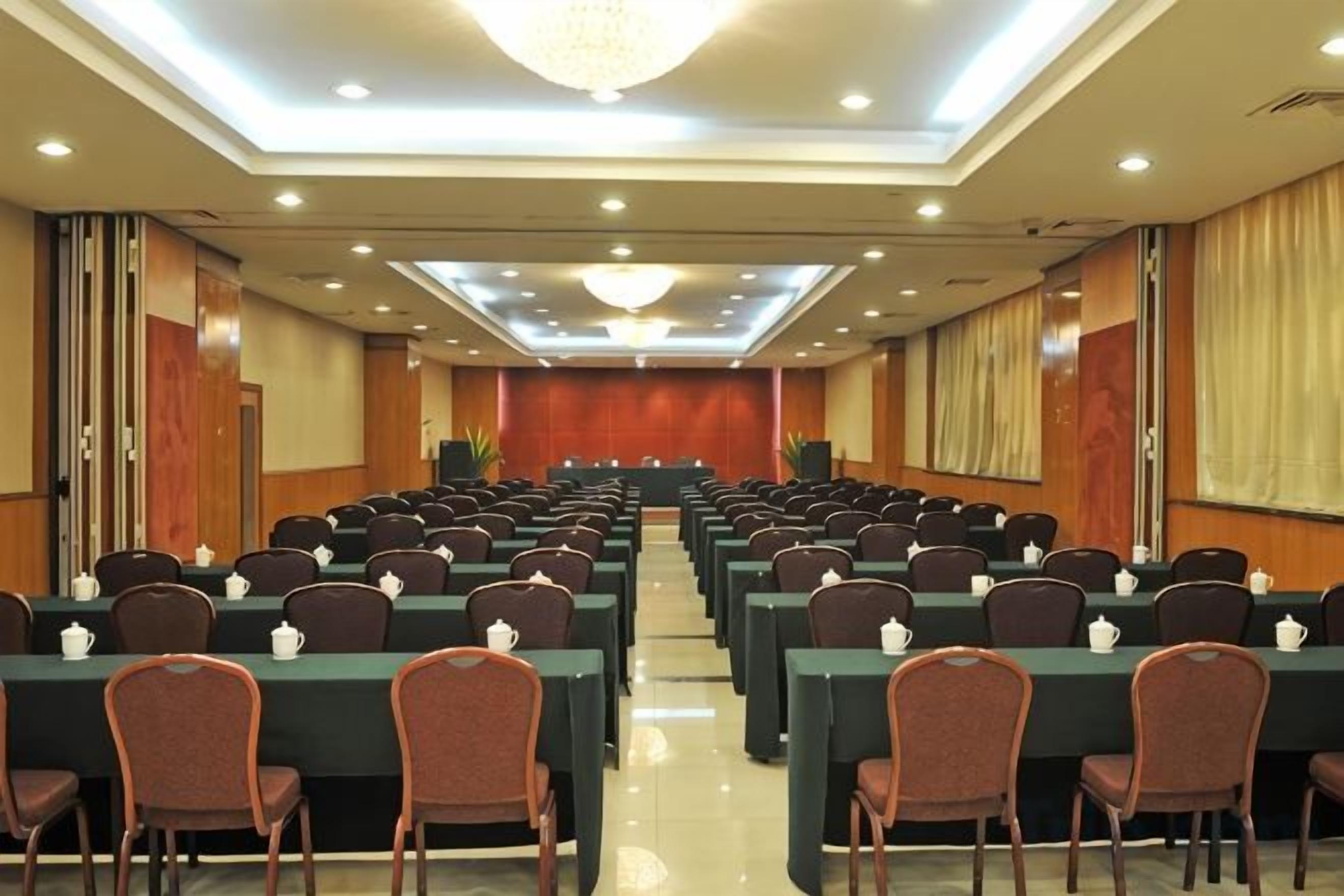 Une salle de conférence avec des rangées de tables et de chaises, une scène à l’avant et un lustre.