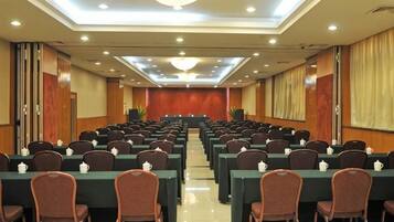 Une salle de conférence avec des rangées de tables et de chaises, une scène à l’avant et un lustre.