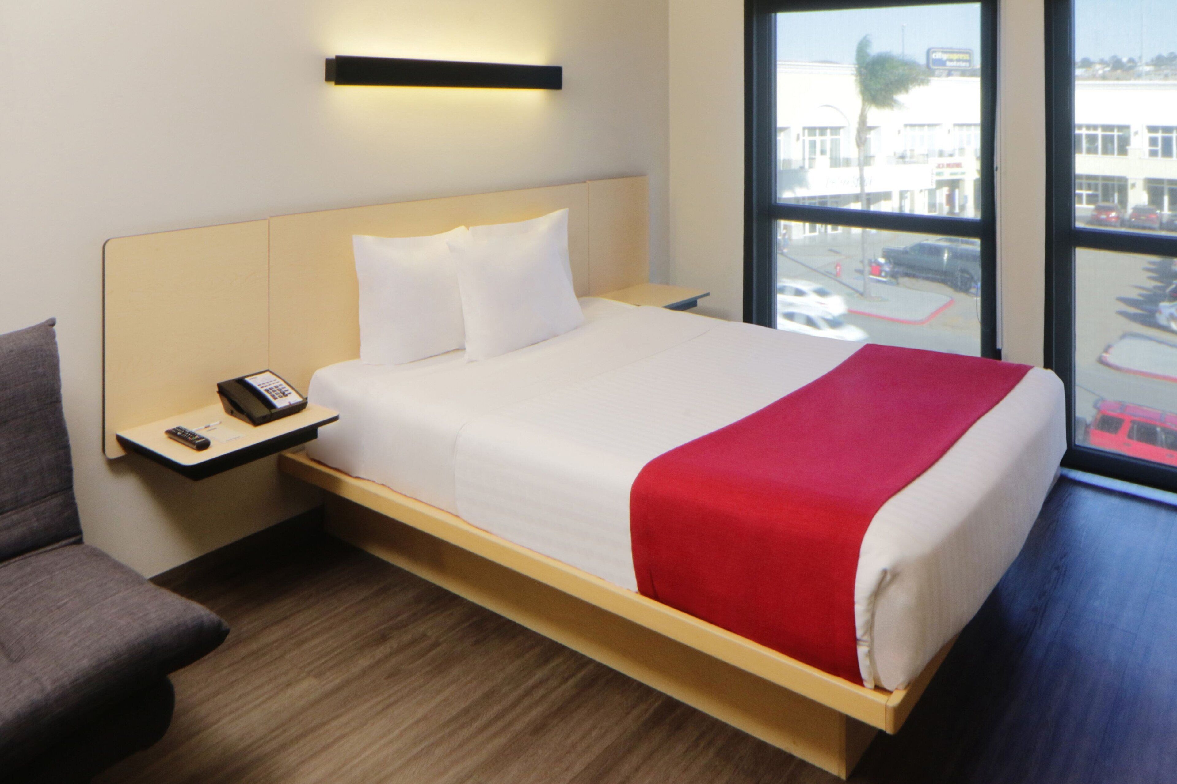 Habitación de hotel con cama, mesita de noche con tel éfono, una silla y un ventanal con vistas a un aparcamiento y edificios.