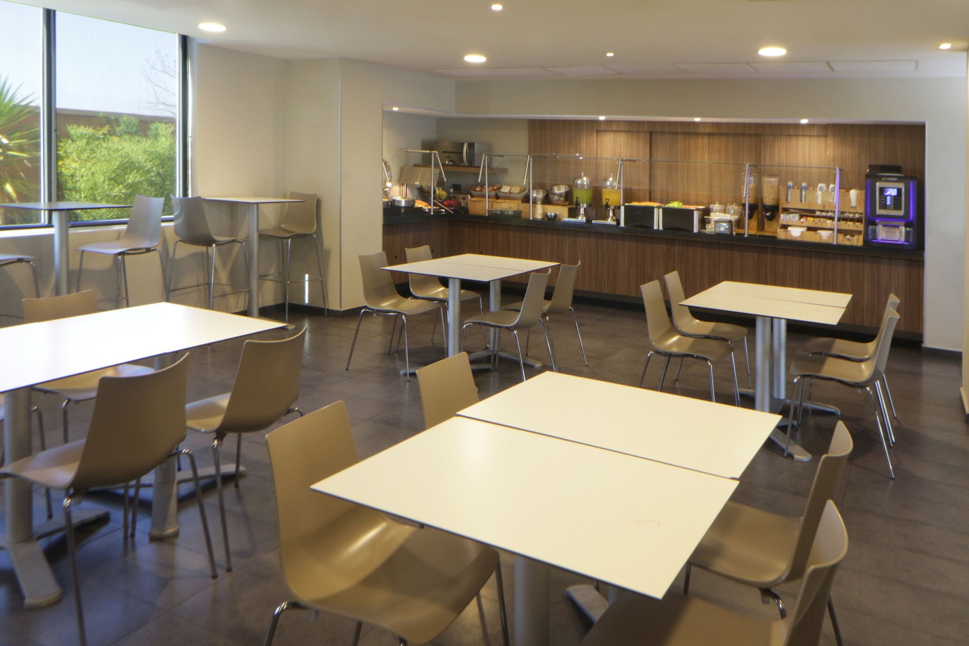 A modern dining area with multiple tables and chairs, a counter with food and beverages, and a view of greenery outside.