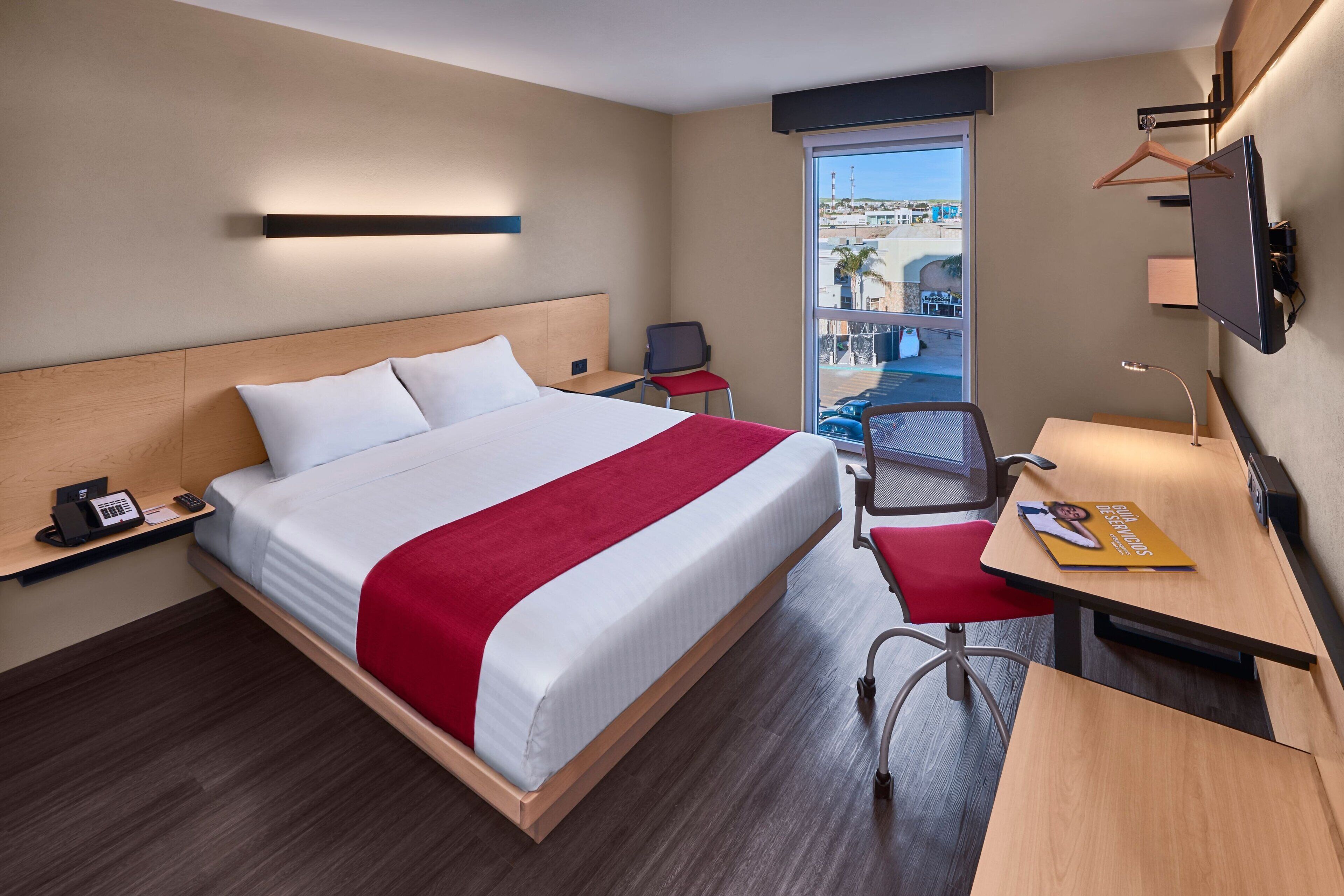 A hotel room with a bed, a desk, a chair, a TV, a phone, and a window with a view of the outside.