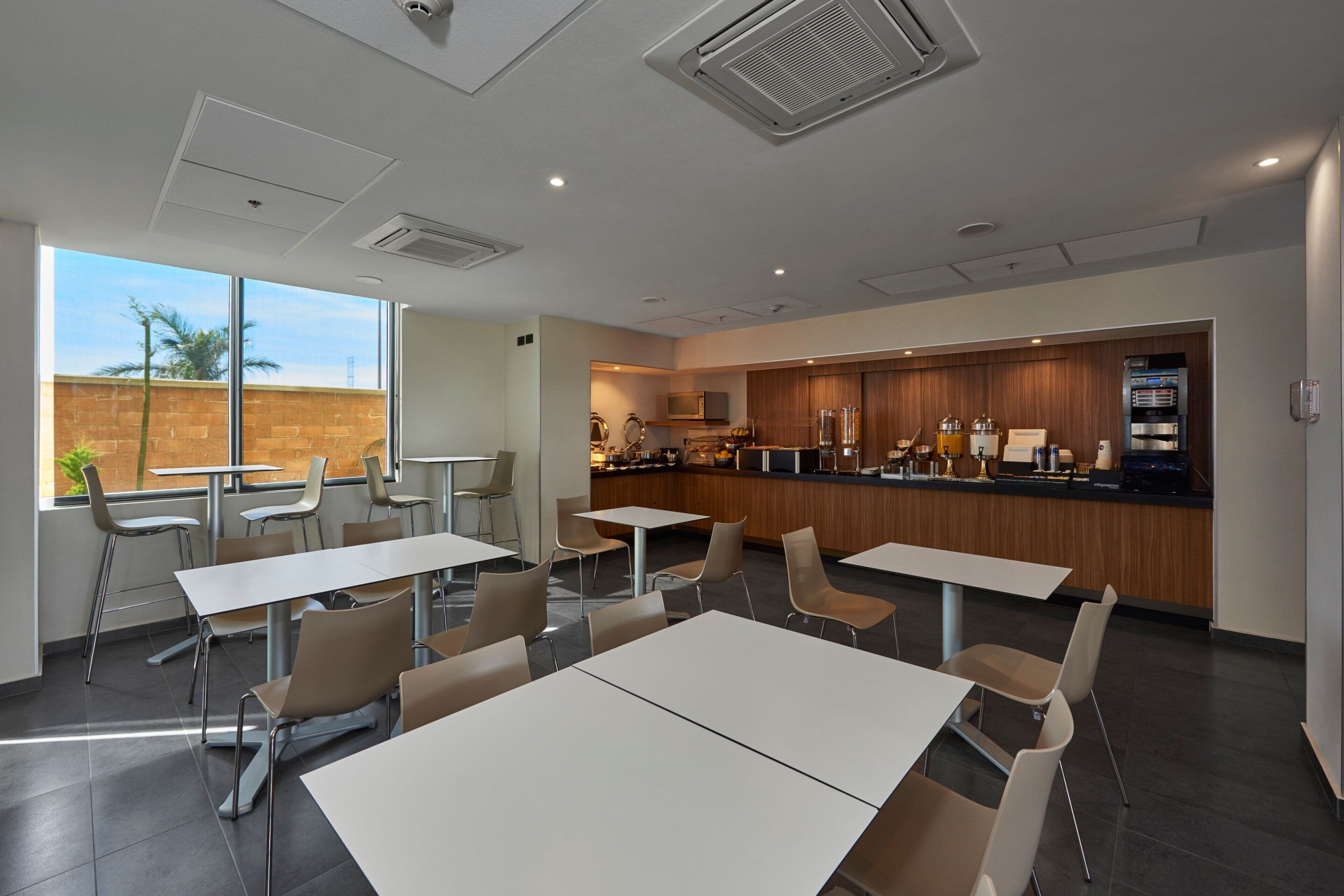 A modern dining area with tables and chairs, a kitchen in the background, and a view of palm trees outside.