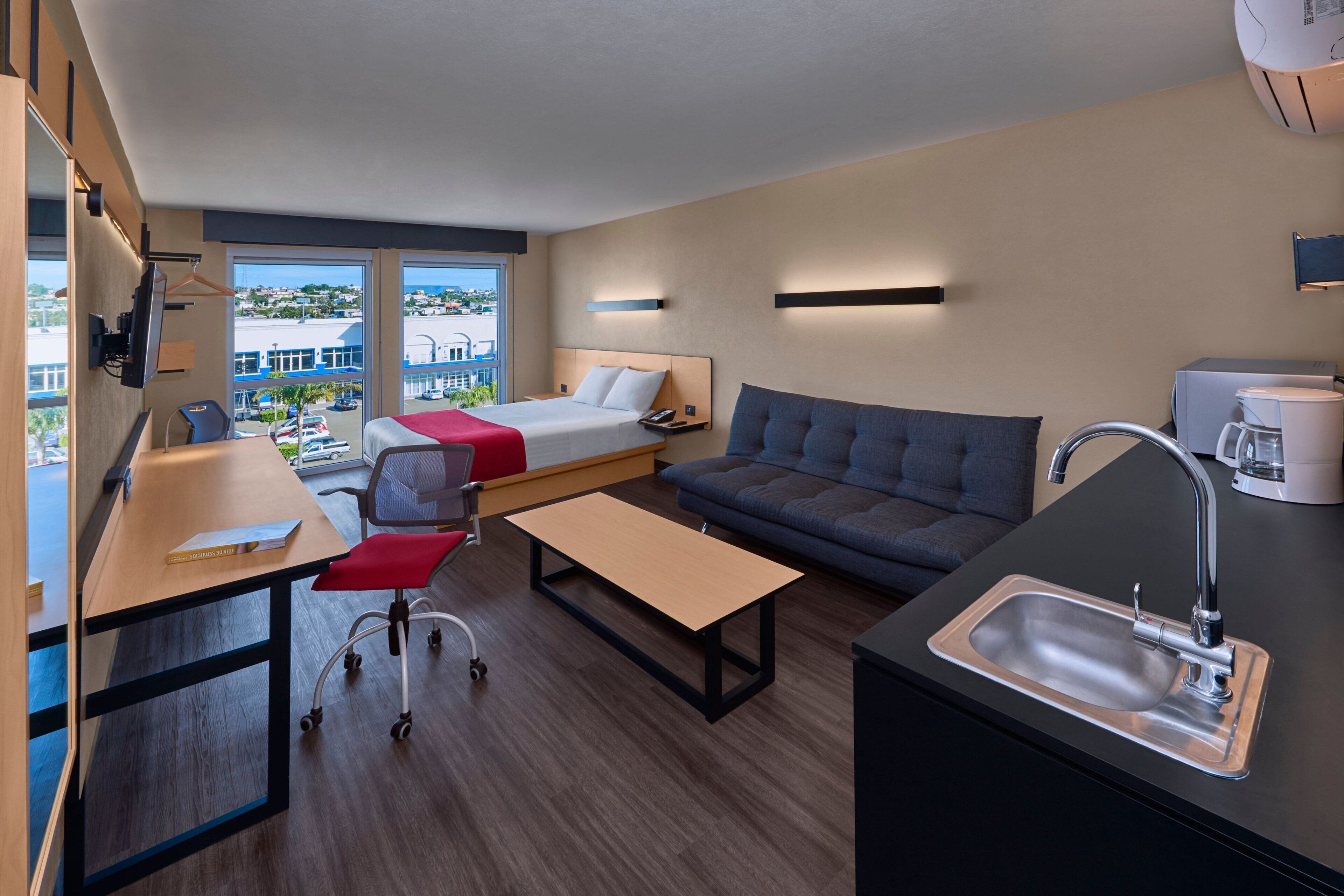 A modern hotel room with a bed, sofa, desk, and kitchenette.